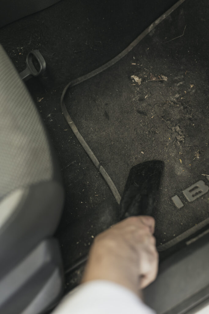 Primer plano de una mano pasando la boquilla de un aspirador sobre una alfombrilla negra con suciedad en el suelo de un coche en Gasolinera Teckmar