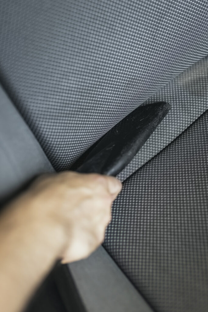 Primer plano de una mano utilizando la boquilla estrecha de un aspirador para limpiar las ranuras de los asientos de tela gris de un coche en Gasolinera Teckmar