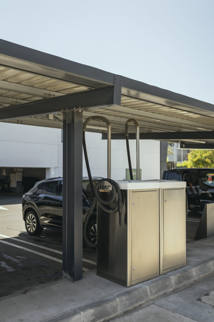 Vista lateral de dos máquinas de aspirado y limpieza de vehículos de acero inoxidable bajo una estructura metálica techada en Gasolinera Teckmar, con un coche negro estacionado al fondo