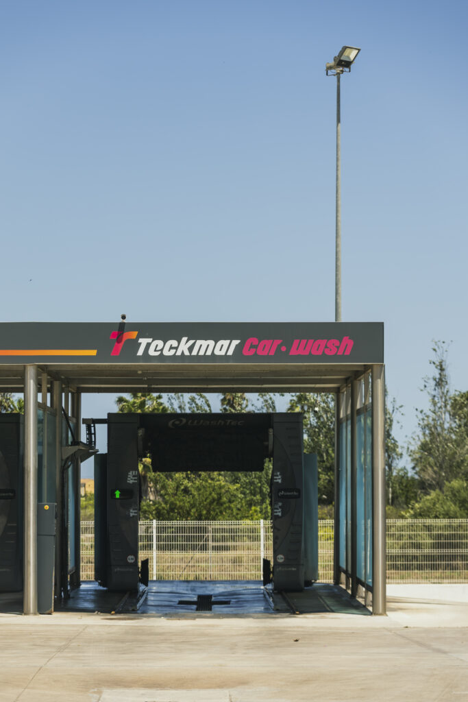Vista frontal de la estructura del túnel de lavado automático WashTec en Teckmar Car Wash, sin vehículos en su interior y con un gran poste de luz de fondo bajo el cielo azul